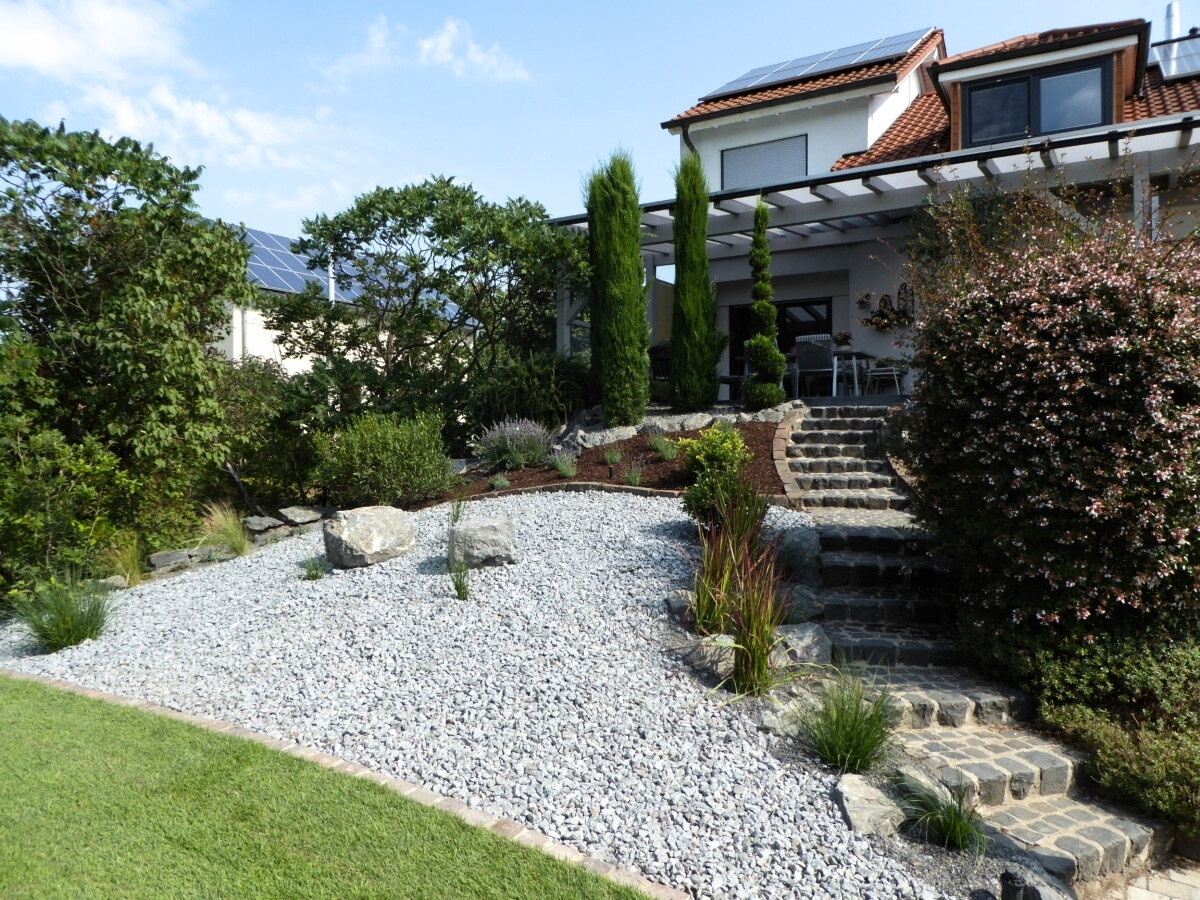 Zweistöckiges Haus mit rotem Ziegeldach, umgeben von Garten mit Rasenfläche, Kiesweg und verschiedenen Büschen und Bäumen.