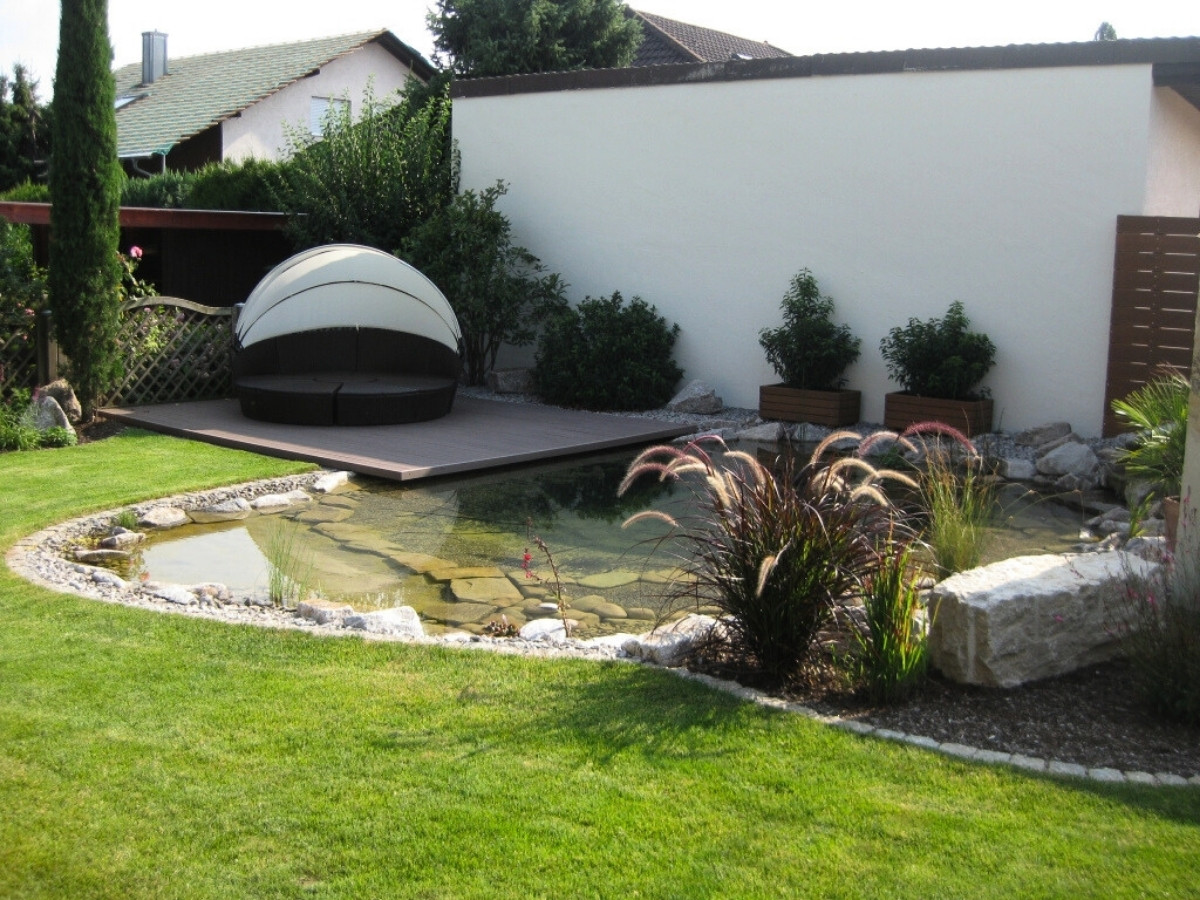 Garten mit kleinem Teich, umgeben von Rasen, Pflanzen und einer runden, halb geschlossenen Gartenliege auf einer Holzterrasse.